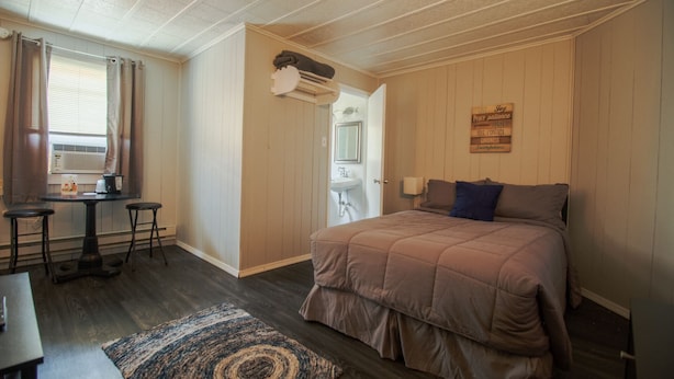 A bedroom with a bed, a desk, and a chair. There is a window with curtains, a radiator, and a wall-mounted shelf with a towel. - Current River Inn (Van Buren)