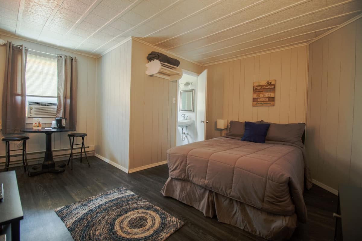 A bedroom with a bed, a desk, and a chair. There is a window with curtains, a radiator, and a wall-mounted shelf with a towel.