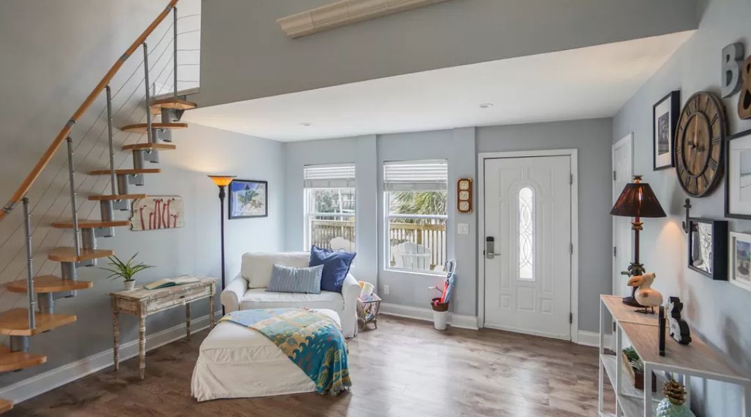 A spacious room with a wooden staircase, a bed with a colorful blanket, a white sofa, and a small table with a lamp.