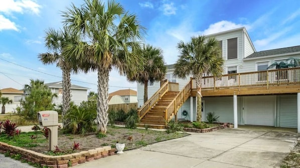 A residential house with a wooden staircase leading to a balcony, surrounded by palm trees and a well-maintained garden.