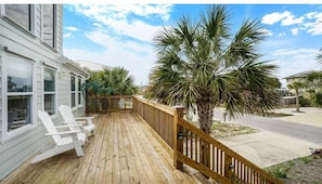 A wooden deck with white Adirondack chairs, a palm tree, and a house with a white exterior.