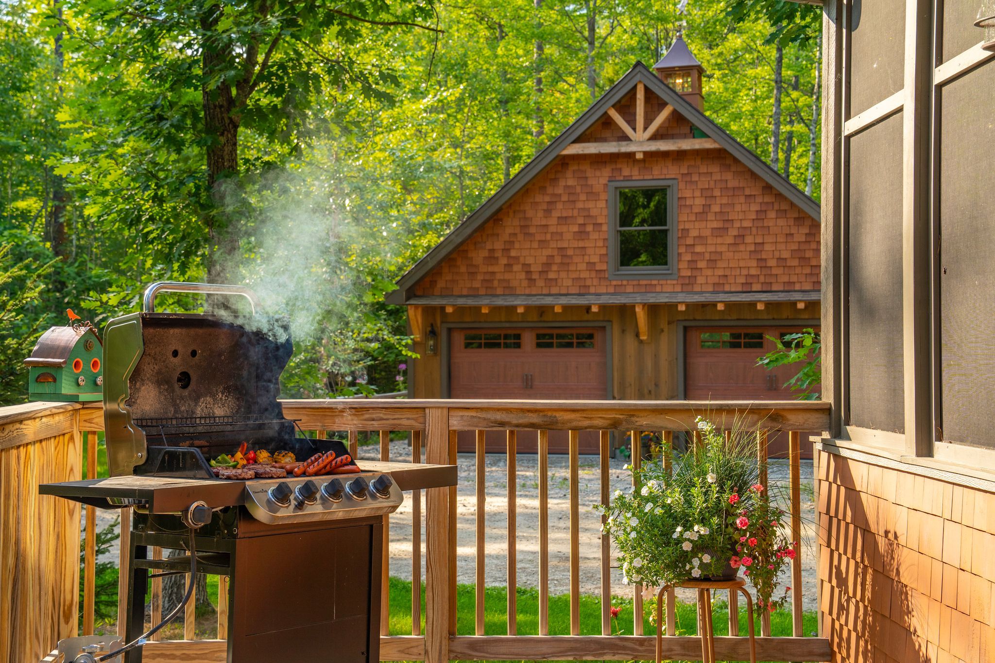 An outdoor grill with food, a wooden deck, and a house in the background.