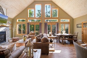 A spacious living room with a stone fireplace, comfortable seating, a dining area, and large windows offering a view of the outdoors.