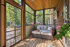 A covered porch with a wooden bench, a wicker ottoman, and a view of the surrounding forest.