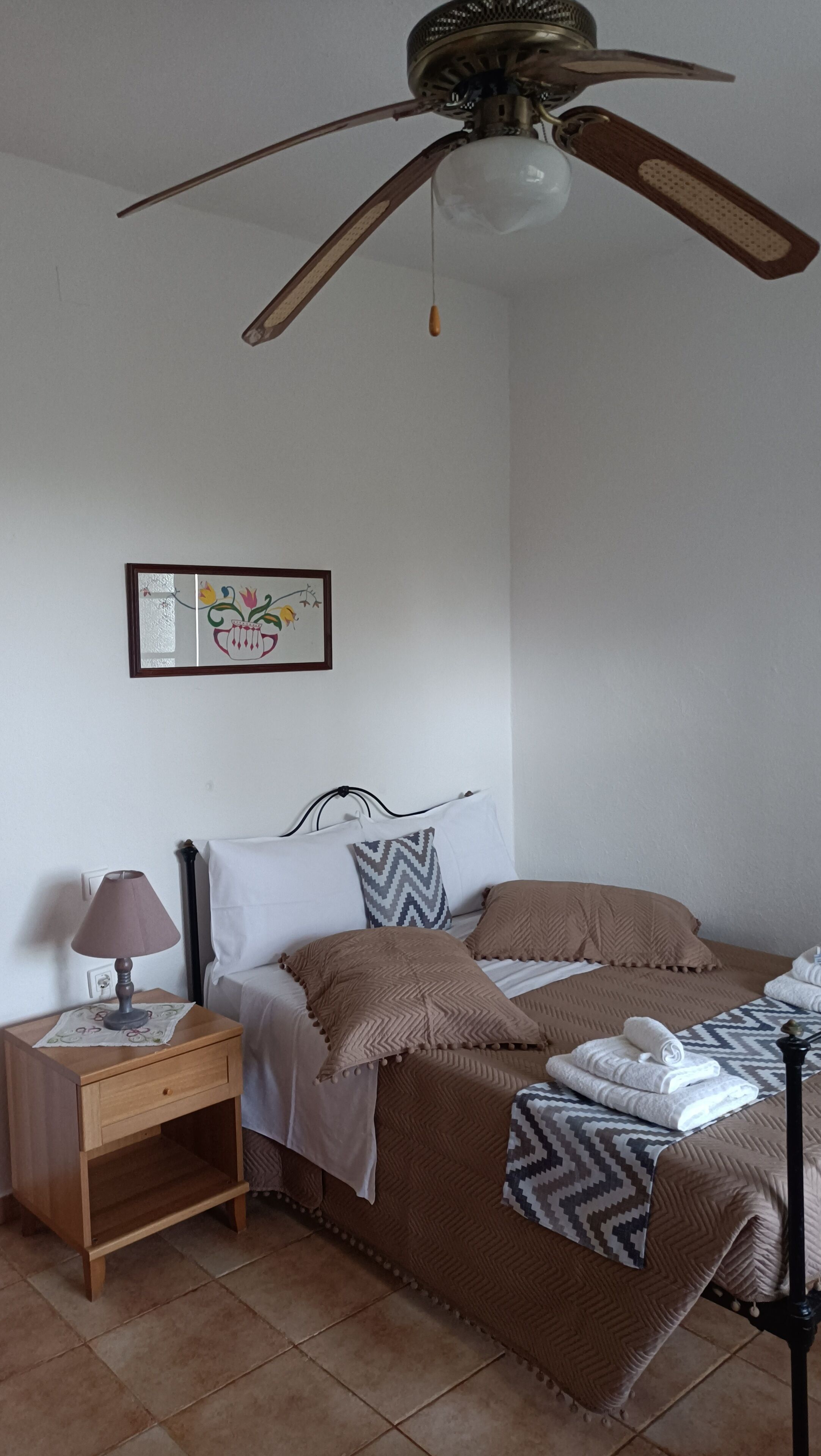 Une chambre à coucher comprenant un lit, une table de chevet, un ventilateur de plafond et un tableau accroché au mur.