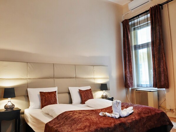 A hotel room with a large bed, two bedside tables, a nightstand, a window with curtains, and a wall-mounted air conditioner. - Folk Art Apartment (Budapest)