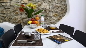 Une table à manger avec un mur en pierre en arrière-plan, un saladier de fruits, une bouteille de jus, des croissants et un magazine.