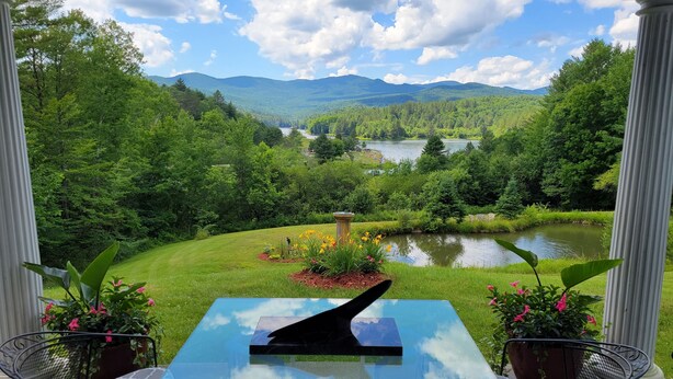 Un patio con una mesa y sillas de vidrio, con vista a un paisaje exuberante con un lago y montañas.