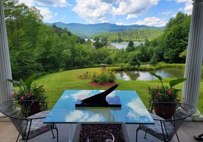 A patio with a glass table and chairs, overlooking a lush landscape with a lake and mountains. - Sir Richards Waterbury Stowe Lake View Escape - ski - kayak -  privacy (Waterbury Center)