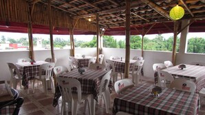 An outdoor restaurant with tables covered in plaid tablecloths and white plastic chairs, featuring a bamboo ceiling and a view of greenery. - Hotel Siesta (Ixtaczoquitlán)
