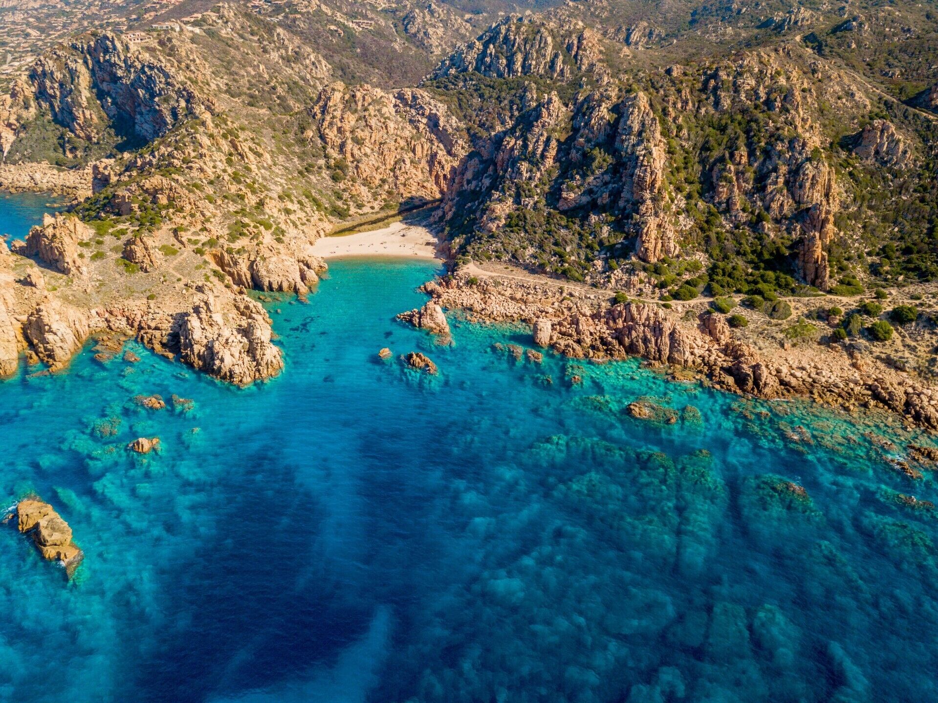 Vue aérienne d’une zone côtière caractérisée par un terrain rocheux, des eaux turquoise et limpides, et une plage de sable.