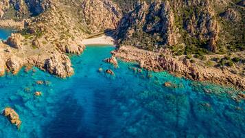 Vue aérienne d’une zone côtière caractérisée par un terrain rocheux, des eaux turquoise et limpides, et une plage de sable.
