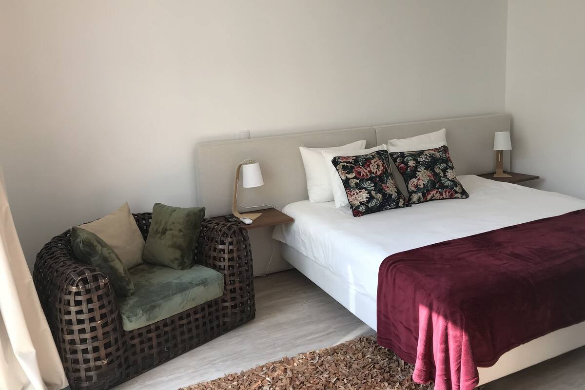 A bedroom with a bed, a nightstand, a lamp, a wicker chair, and a blanket.
