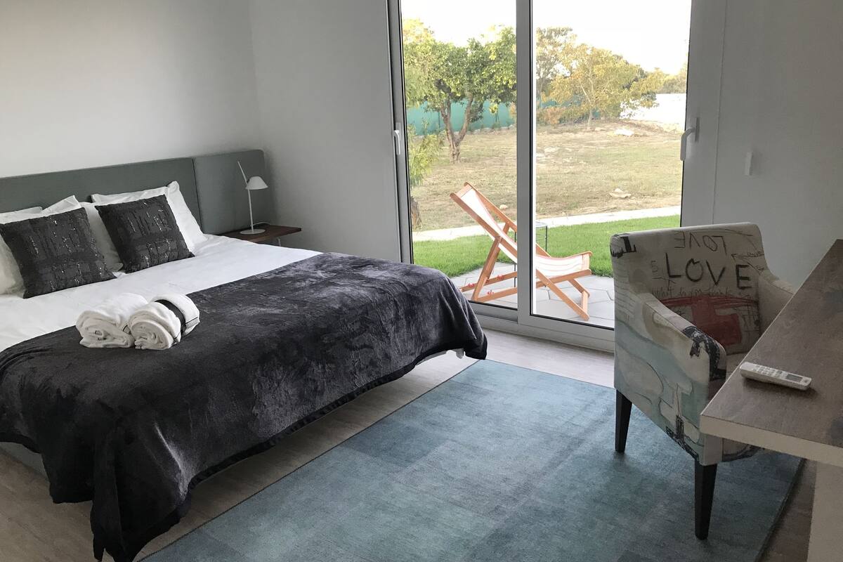 A bedroom with a bed, a nightstand, a lamp, a window, and a view of the outdoors.