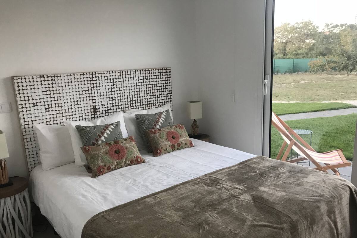 A bedroom with a large bed, multiple pillows, a headboard with a circular pattern, a bedside table with a lamp, and a view of a grassy area outside.
