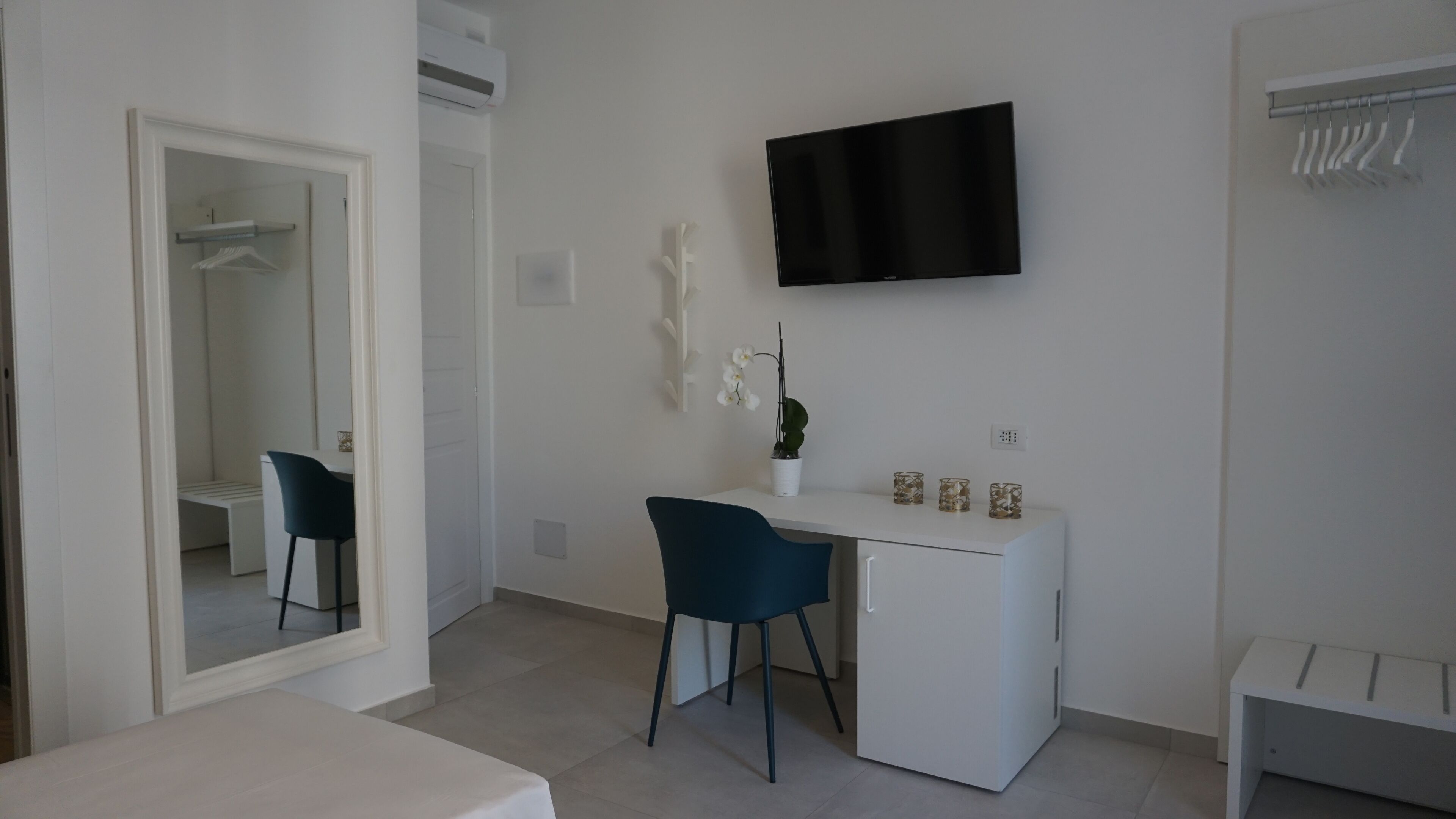 Une pièce moderne et minimaliste, avec un bureau blanc, une chaise, un téléviseur à écran plat fixé au mur et une petite orchidée en pot posée sur le bureau.