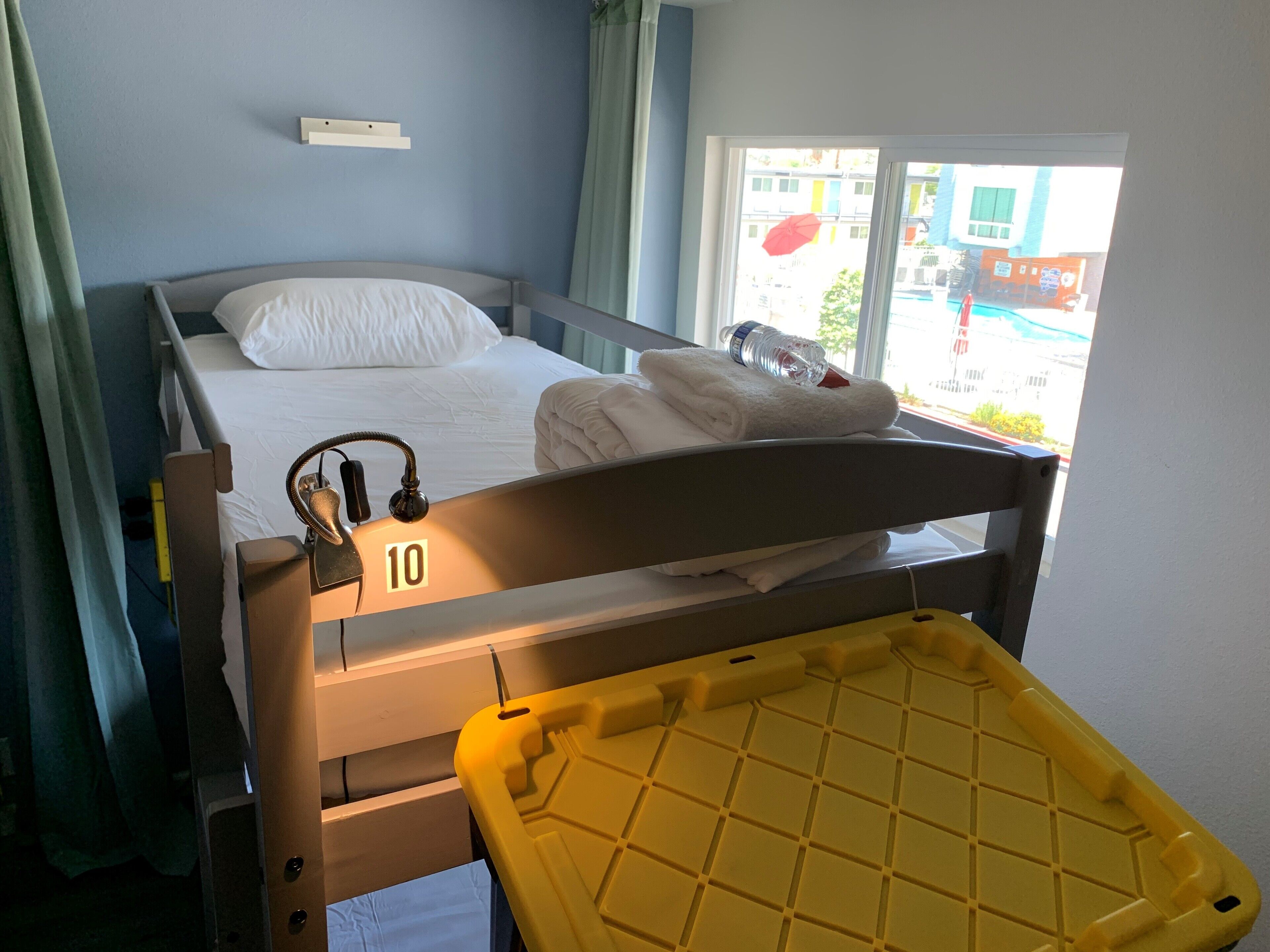 A single bed with a number '10' on the headboard, a bedside table with a lamp, and a window with a view of the outside.