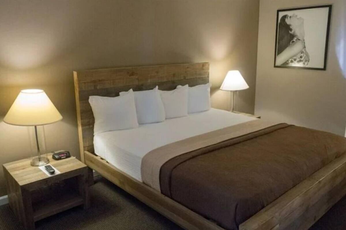 A single bed with a wooden headboard, two bedside lamps, a remote control on the nightstand, and a framed picture on the wall.