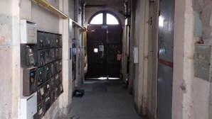 A narrow corridor with a high ceiling, a large arched window at the end, and a door with a sign.
