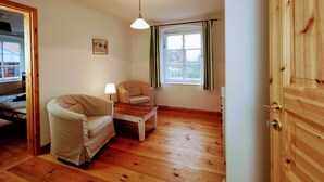 Een kamer met een houten vloer, een bank, een bijzettafeltje, een lamp, een raam met gordijnen en een deur.