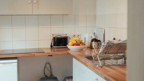 Een kleine keuken met een houten aanrecht, een broodrooster, een fruitschaal en een koffiezetapparaat. Er staan twee prullenbakken en er hangt een lamp aan de muur.