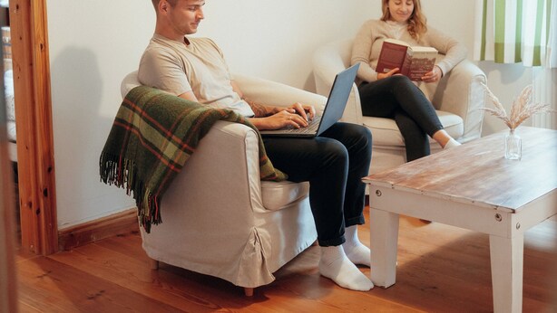 Een kamer met een houten vloer, een witte bank, een bijzettafeltje en een persoon die een laptop gebruikt.