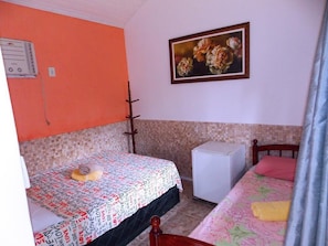 A small, single-bed room with a tiled floor, a bed with a patterned bedspread, a small white cabinet, and a painting on the wall. - Pousada L'Auberge (Angra dos Reis)