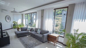 A modern living room with a grey sectional sofa, a wooden side table, a large window with curtains, and a potted plant. - El Cubil 8 (Sardinal)
