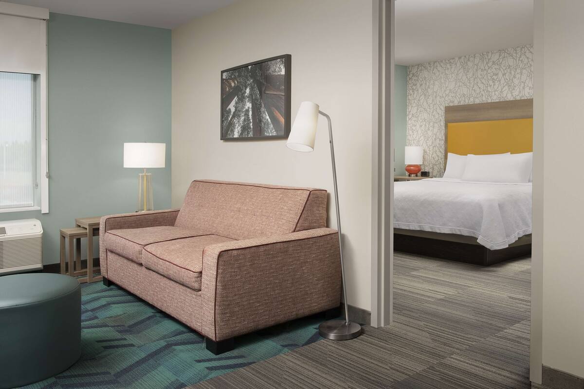 A hotel room with a sofa, a bedside table with a lamp, a bed with a headboard, and a wall with a framed picture.