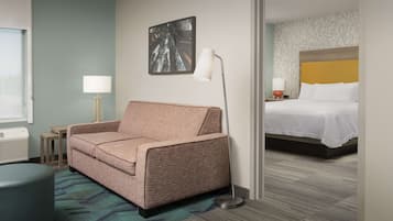 A hotel room with a sofa, a bedside table with a lamp, a bed with a headboard, and a wall with a framed picture.