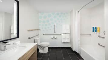 Un baño moderno con lavamanos, inodoro y bañera blancos. Cuenta con un pasamanos, barras de apoyo y un perchero para toallas.