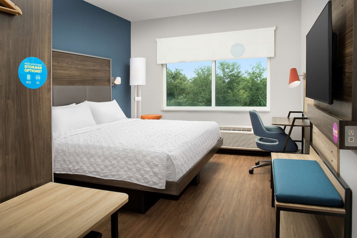 Habitación de hotel moderna con una cama grande, un escritorio, una silla, un televisor y una ventana con vistas a árboles.