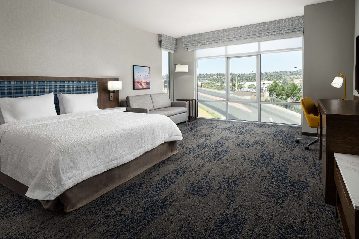 A hotel room with a large bed, a desk, a chair, a sofa, and a large window offering a city view.