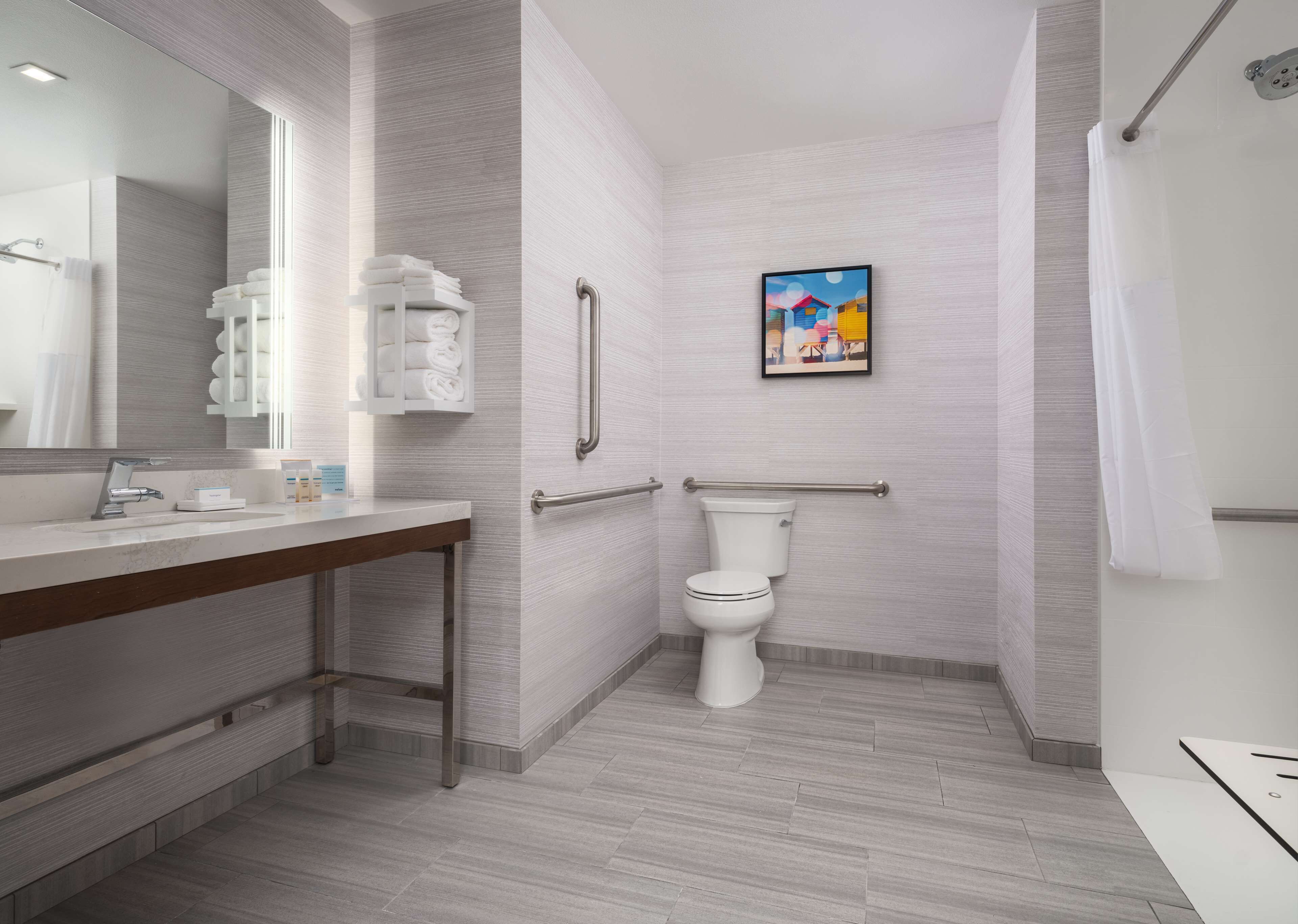 Un baño moderno con lavamanos, inodoro y ducha blancos. Se observan barras de apoyo y un perchero para toallas.