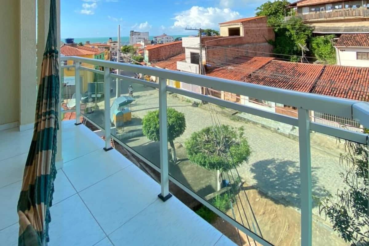 Ein Balkon mit Blick auf eine Wohngegend, darunter Häuser mit roten Ziegeldächern und einem klaren, blauen Himmel.