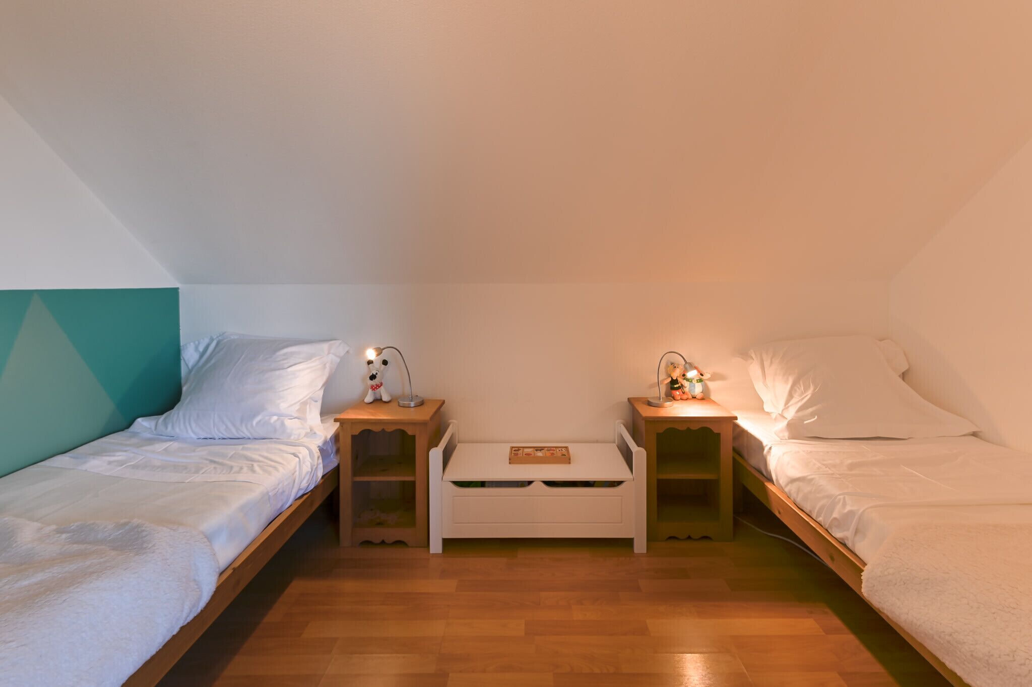 A small, cozy room with two single beds, each with a nightstand and a lamp. There is a white storage box in the center of the room.