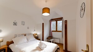 A neatly made bed with white linens and towels, a bedside table with a lamp, a small wooden stool, and a window with curtains. - The green fee, close to the Golf course and 5 minutes from the lake (Giez)
