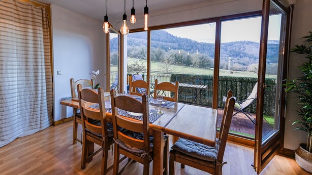 A dining area with a wooden table and chairs, large windows with a view of a green landscape, and hanging light fixtures. - The green fee, close to the Golf course and 5 minutes from the lake (Giez)