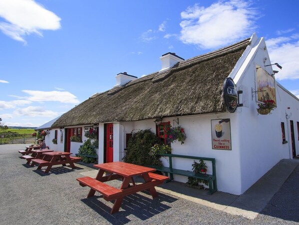 Un bâtiment blanc avec un toit de chaume, une porte rouge et une enseigne publicitaire pour la bière Guinness. Il y a des tables de pique-nique à l'extérieur.