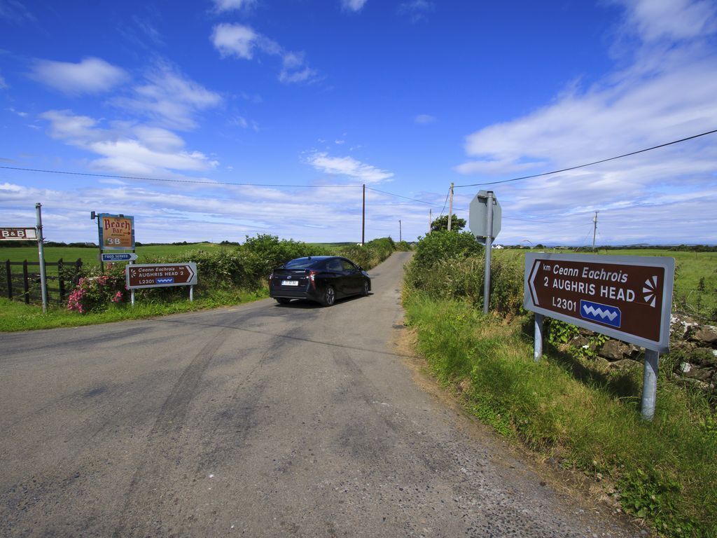 Uma estrada rural com placas de sinalização, um carro e o anúncio de um bar de praia.