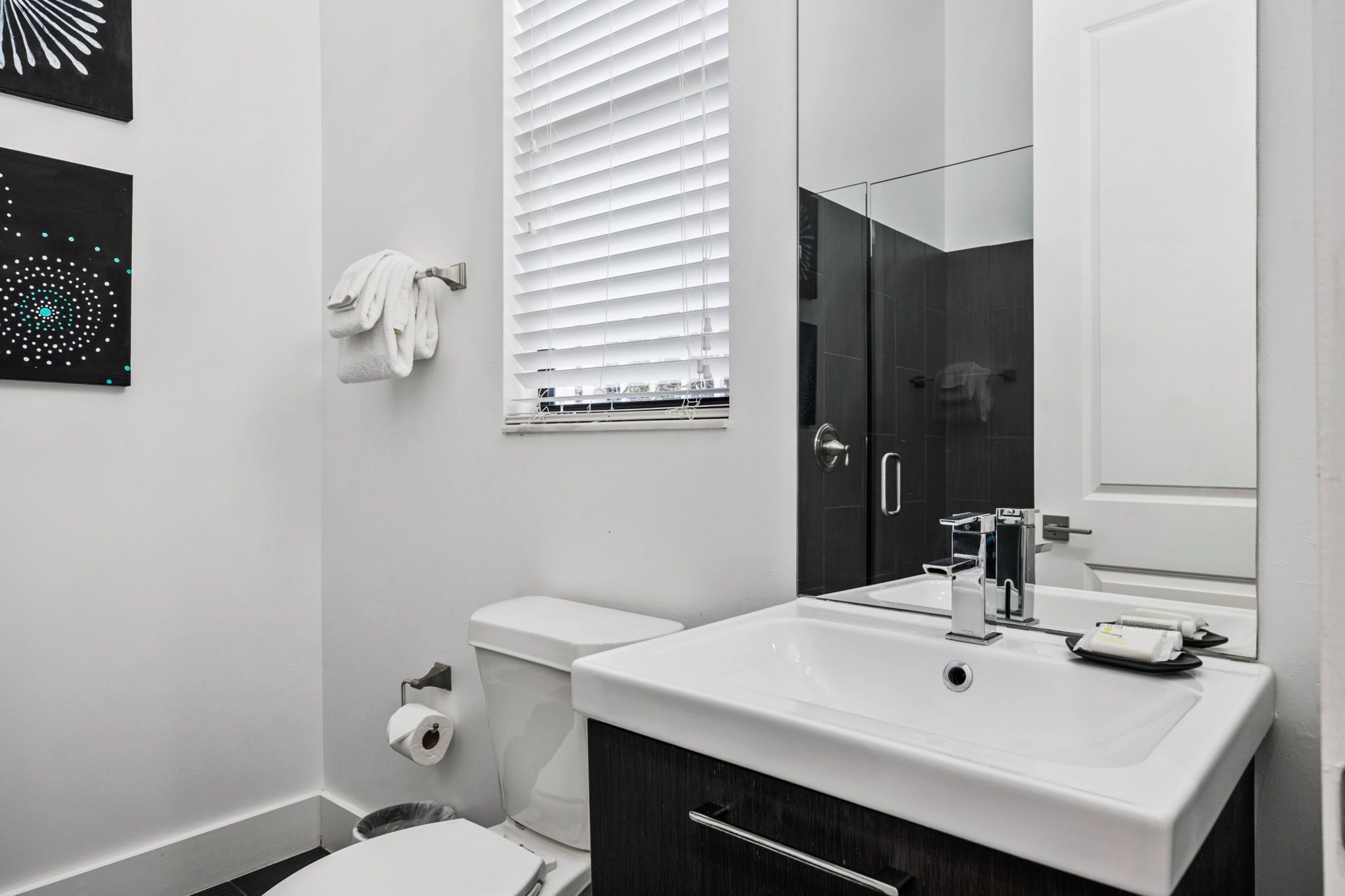 Ein modernes Badezimmer mit einem weißen Waschbecken, dunklen Schränken, einer Toilette und einem Fenster mit Jalousien.
