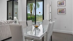 A modern dining area with a glass table, white chairs, and a view of a garden through glass doors. - Liberty Holiday: Private Pool, Water Park, Golf View, Near Disney (Kissimmee)
