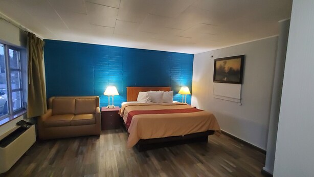 A hotel room with a bed, a sofa, two bedside lamps, a window with curtains, and a painting on the wall. - Blue Haven Motel I-75 North Exit 128&129 (Rocky Top)