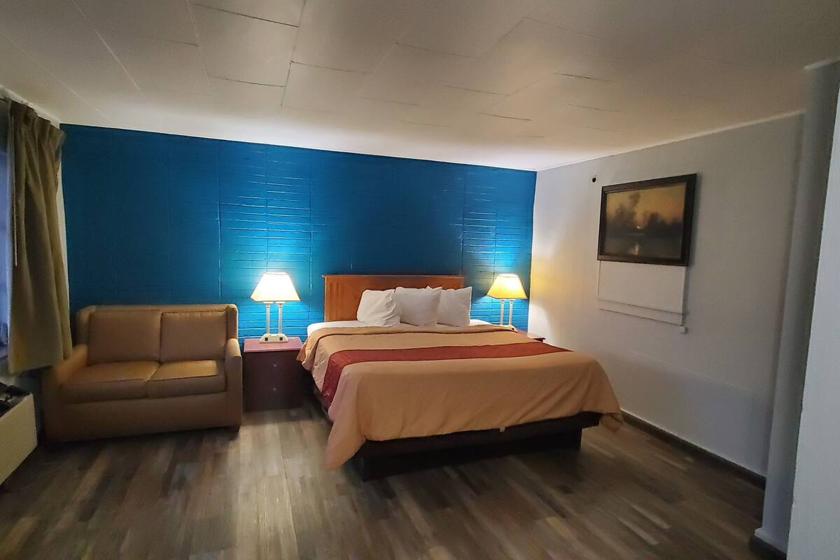 A hotel room with a bed, a sofa, two bedside lamps, a window with curtains, and a painting on the wall.