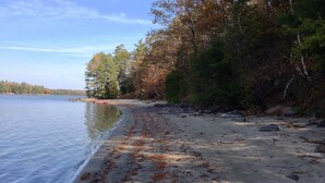 A beach with a sandy shore, scattered rocks, and trees in the background. - Water access Island Cottage in sheltered cove (South Frontenac Region)