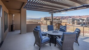Un espace extérieur aménagé, abrité et doté de meubles en osier, avec un foyer central, offrant une vue sur un paysage désertique où l’on aperçoit des bâtiments au loin.