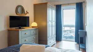 Ein Schlafzimmer mit Bett, Kommode, Fernseher, Lampe und Blick durch ein Fenster.