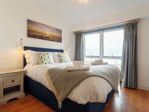A neatly arranged bedroom with a bed, bedside tables, a nightstand, a painting, and a window with curtains. - 2 bedroom accommodation in Newquay (Newquay)