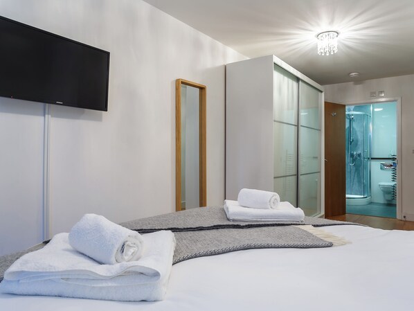A modern hotel room with a bed, a flat-screen TV, a mirror, and a glass shower enclosure.