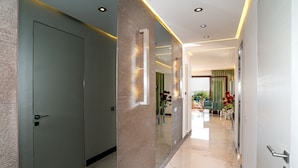 A modern hallway with a mirrored wall, a door, and a view into a room with a green chair and a table.